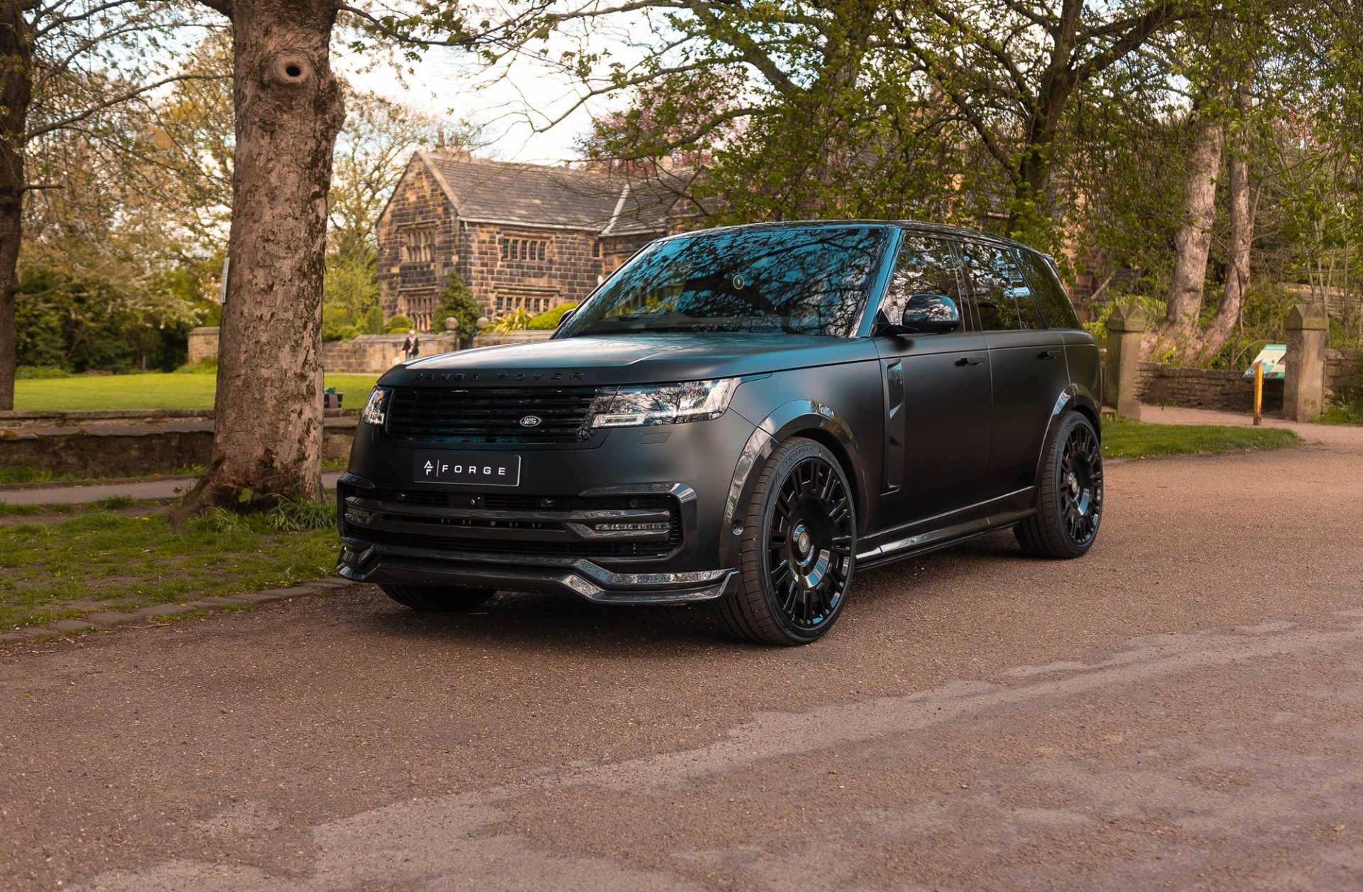 A custom Forge Land Rover Range Rover, matte black, is parked on a gravel path with a historic stone building and trees in the background.