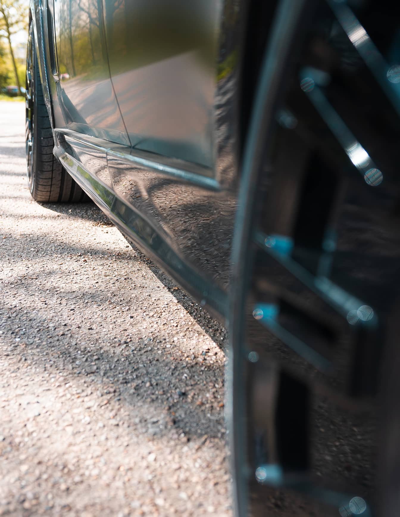A custom Forge Land Rover Range Rover is parked on a gravel surface, with its glossy dark side panel reflecting the sunny outdoor setting.