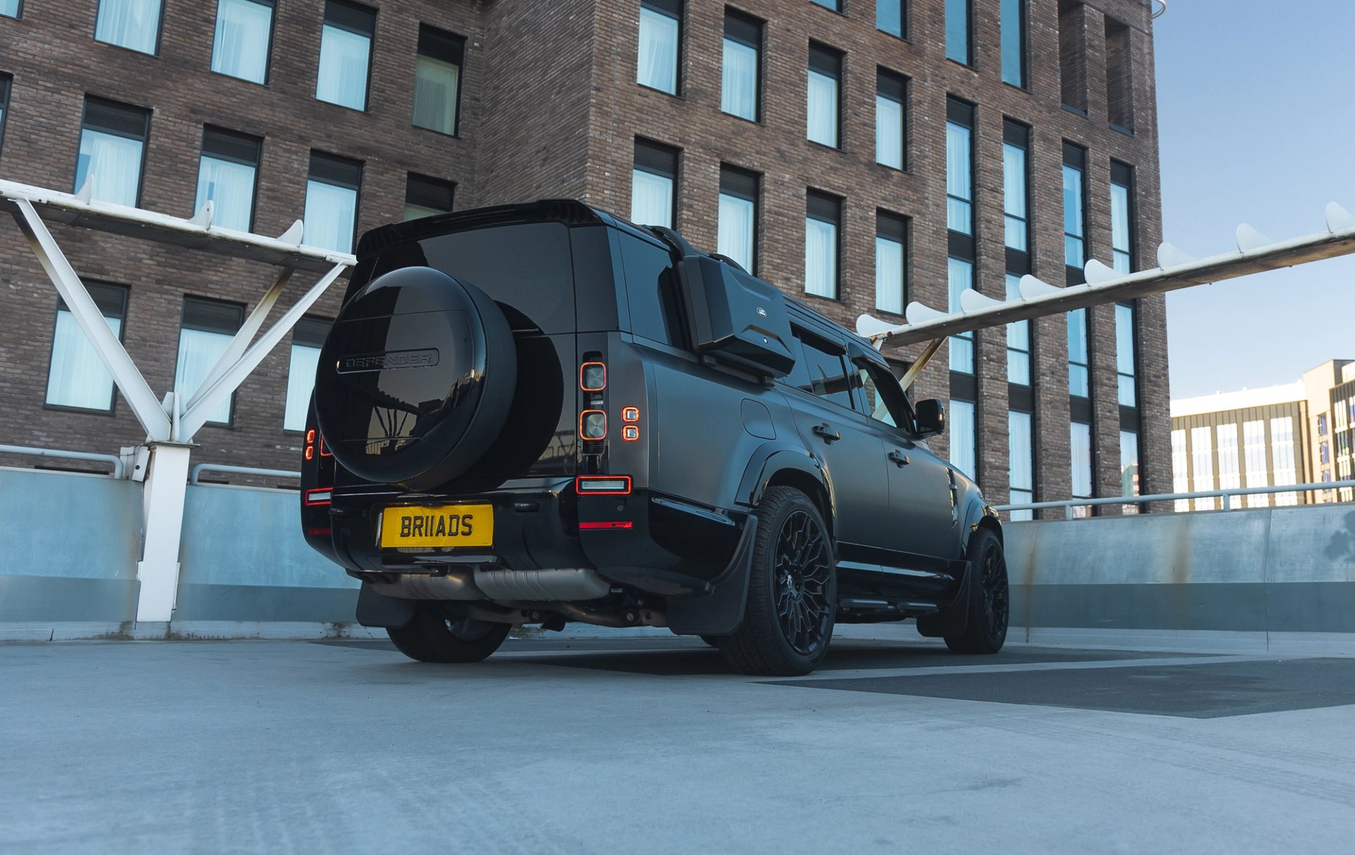 A custom Forge Land Rover Defender 130, matte black, is parked on a concrete rooftop with a brick building in the background.