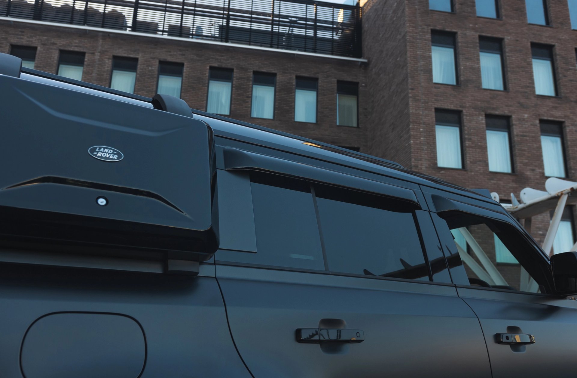 A custom Forge Land Rover Defender 130 with a matte black finish and roof-mounted cargo box is parked in front of a brick building.