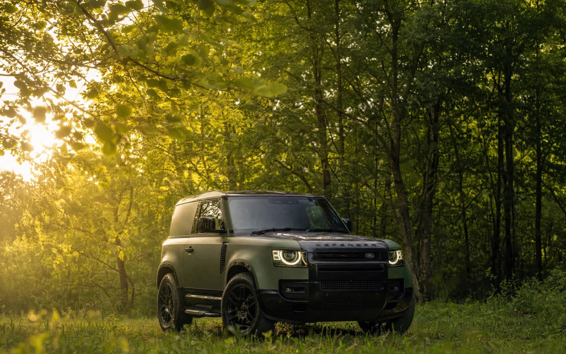 A custom Forge Land Rover Defender 90 parked in a grassy clearing surrounded by lush green trees with sunlight filtering through the leaves.