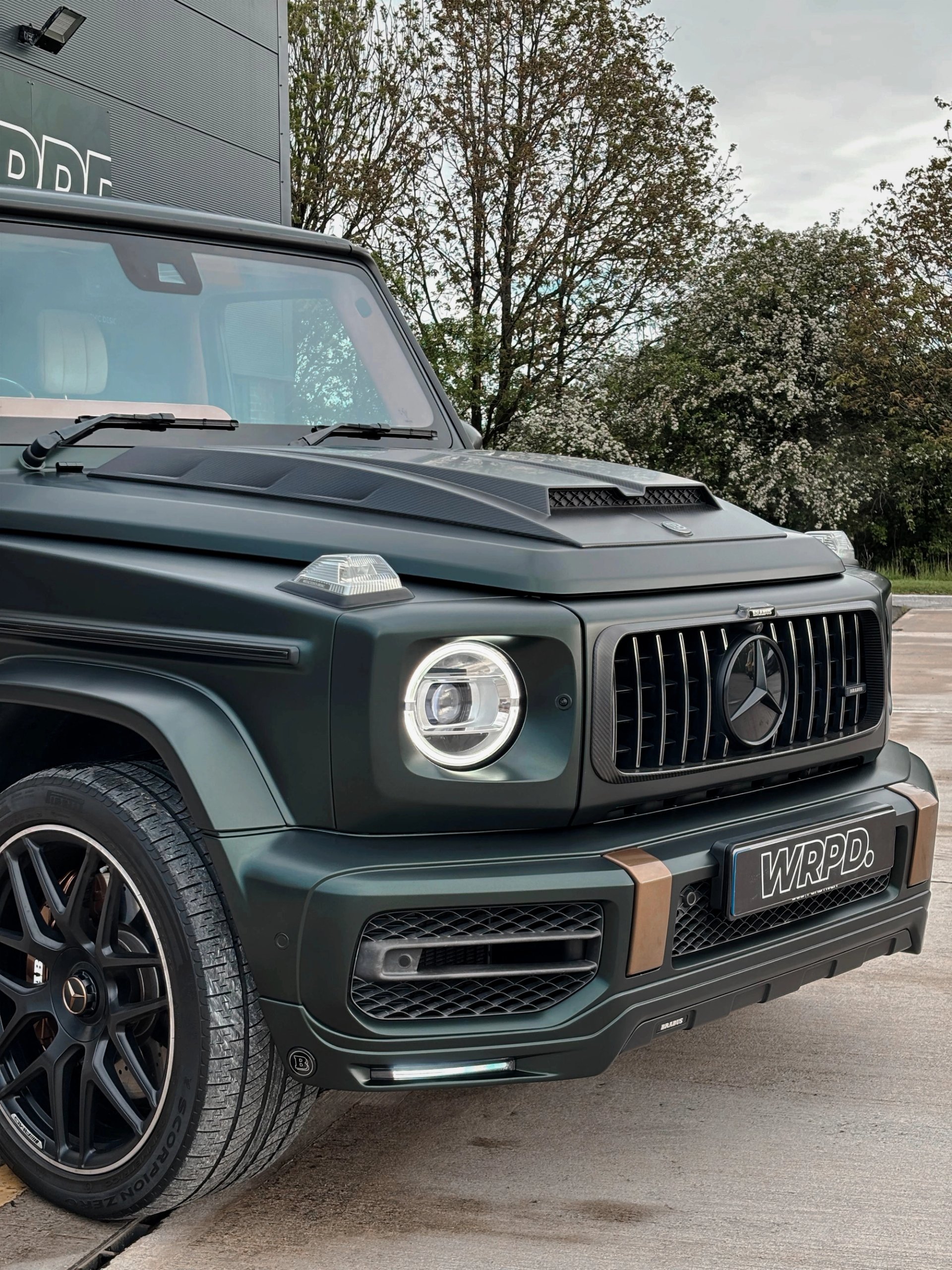 A custom Forge Mercedes G63 with a matte green finish parked on a concrete lot near trees.