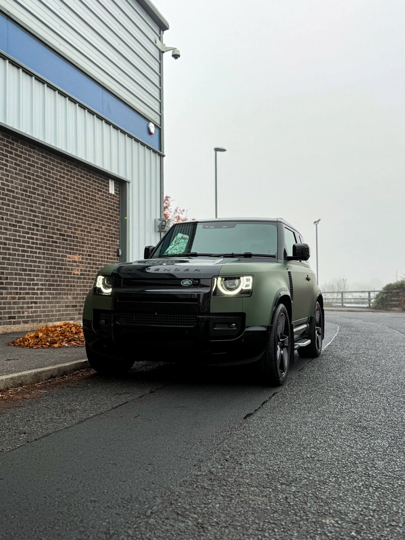 A custom Forge Land Rover Defender 90 parked on an asphalt road beside a brick and metal building on an overcast day.