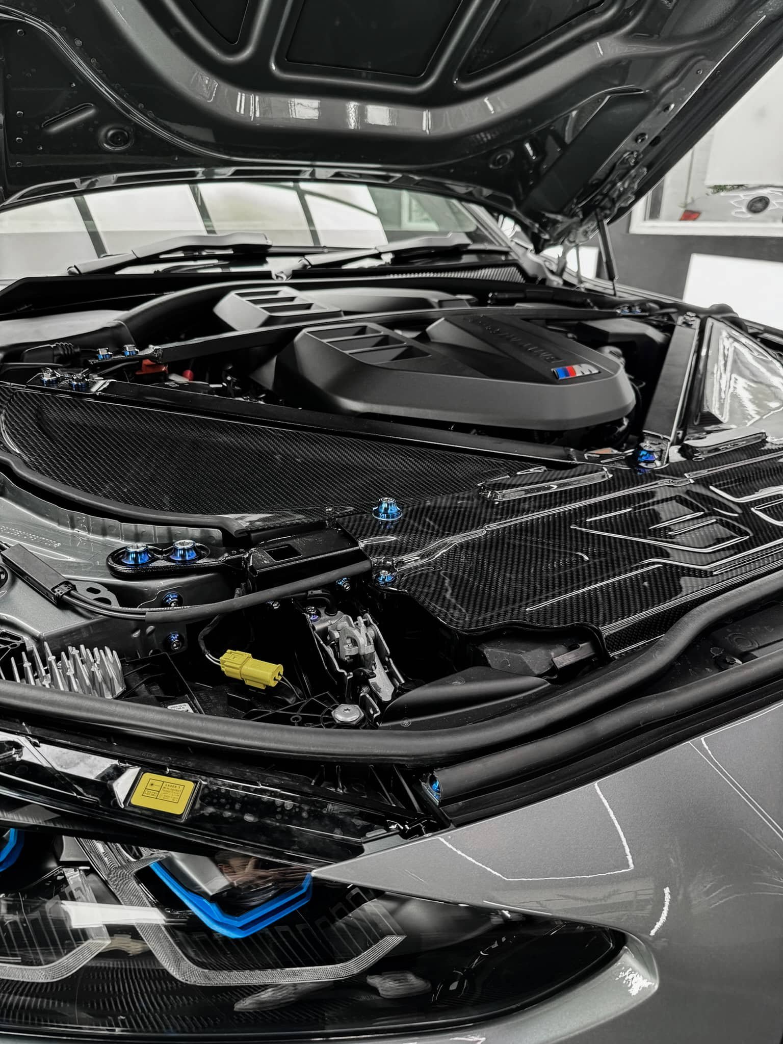 A custom Forge BMW M5 Touring features its open hood revealing a detailed carbon fiber engine bay inside a bright workshop.
