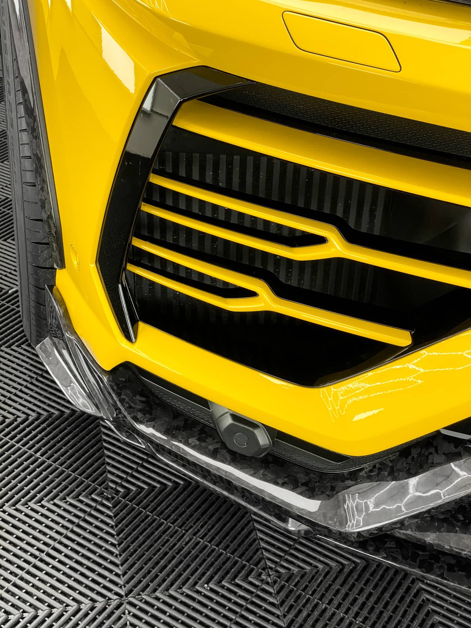 A close-up view shows the yellow front bumper and grille of a custom Forge Lamborghini Urus parked inside a workshop