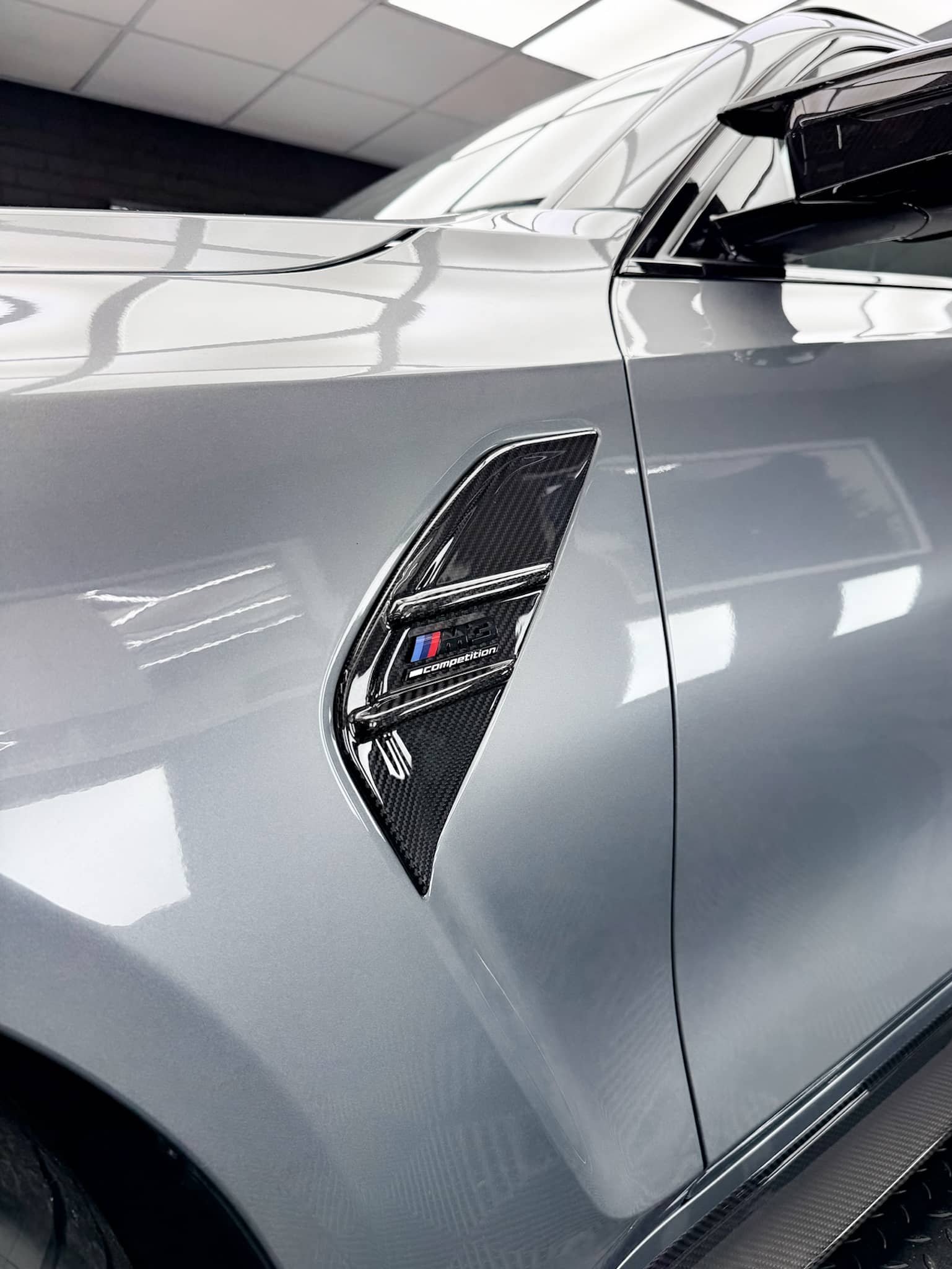 A close-up of a carbon fiber side vent and M5 Competition badge on a silver custom Forge BMW M5 Touring parked inside a brightly lit workshop.