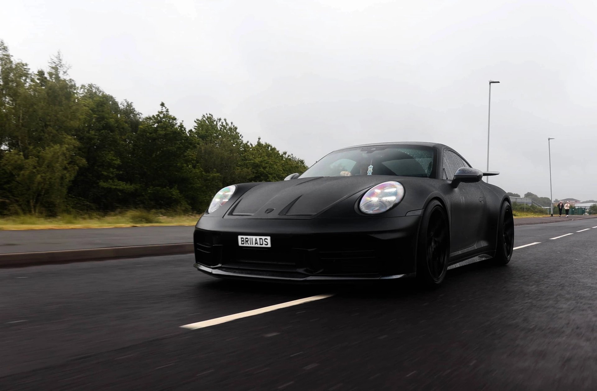A custom Forge Porsche 911 Carrera Turbo in matte black drives on a wet road under an overcast sky, with trees and streetlights in the background.