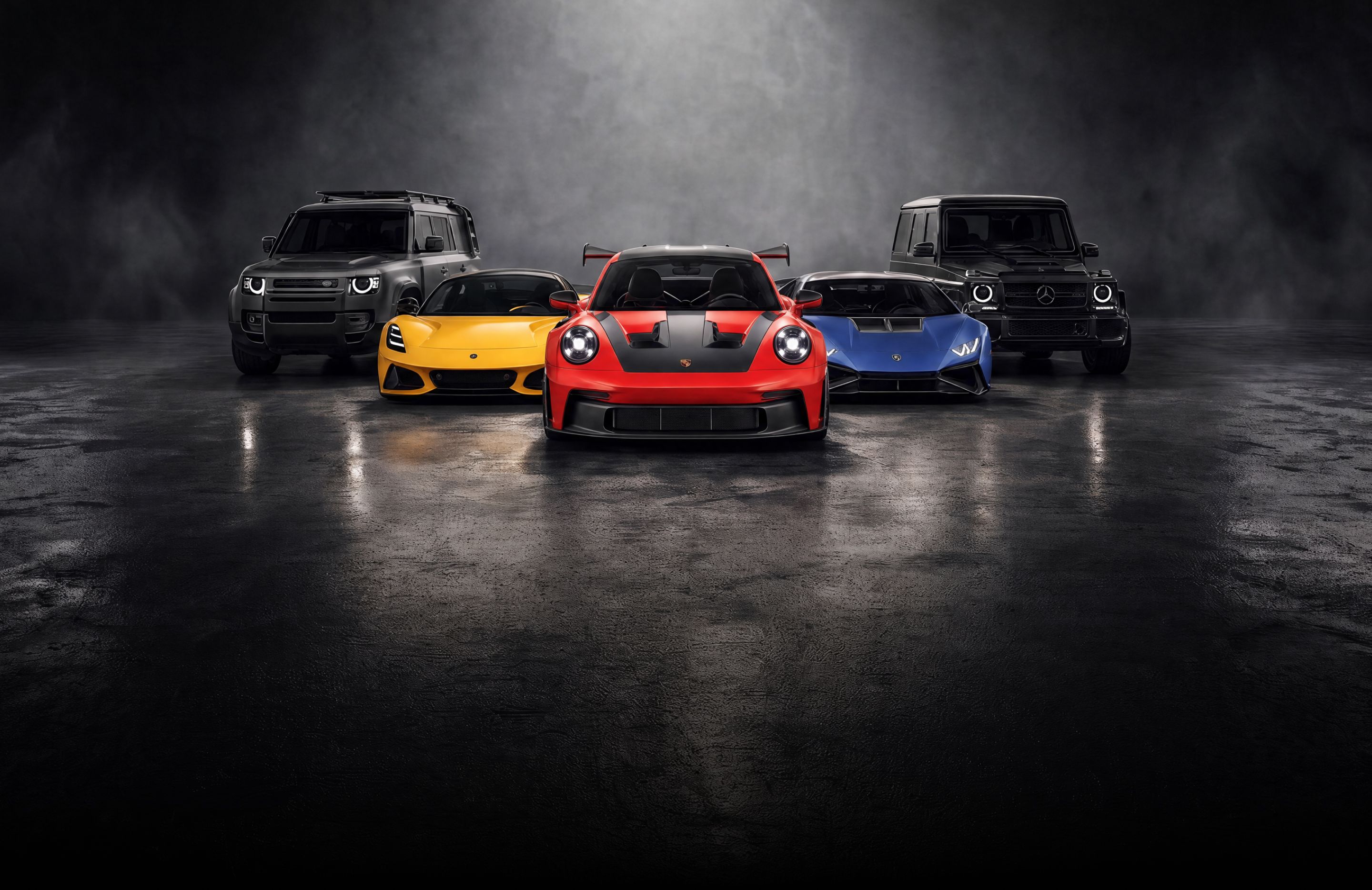 A custom Forge Porsche 911 GT3, Lamborghini Huracan, Lotus Emira, Land Rover Defender, and Mercedes G-Wagen parked on a dark, wet surface.