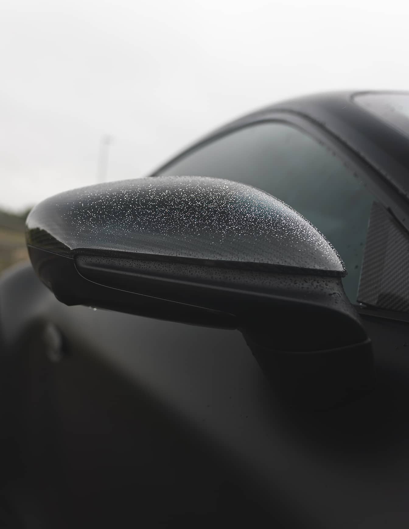 A custom Forge Porsche 911 Carrera Turbo's black side mirror is covered in raindrops on a cloudy day.