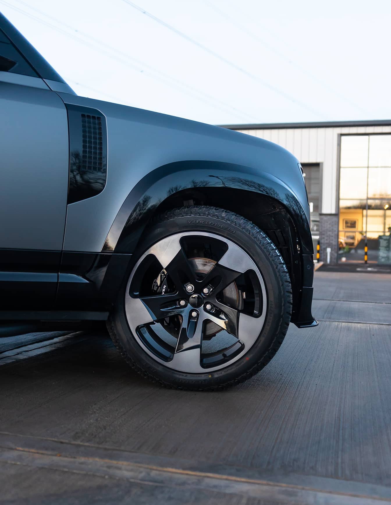 A custom Forge Land Rover Defender 110 is parked on a concrete surface outside a modern building with large windows.