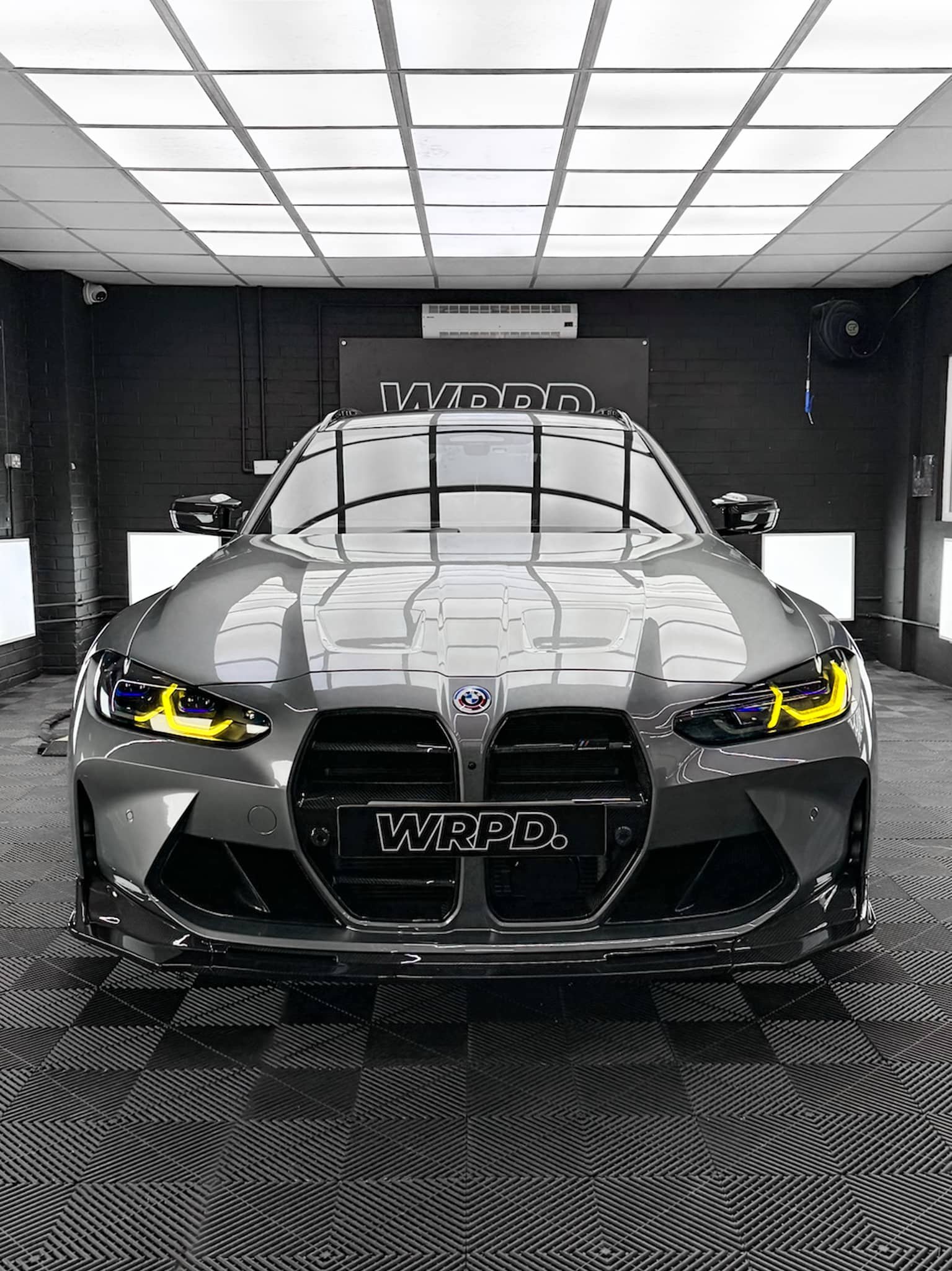 A custom Forge BMW M5 Touring sits centered in a brightly lit, modern black-tiled workshop with overhead square lighting panels.